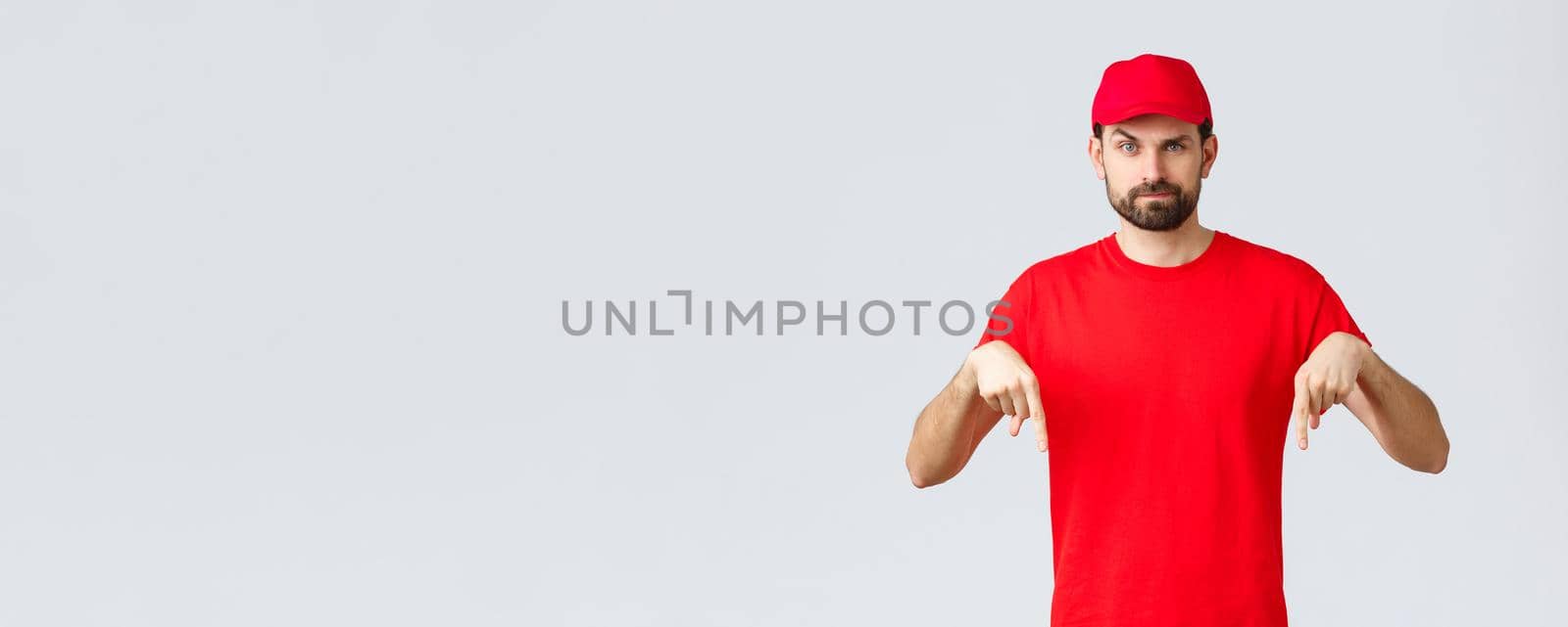 Online shopping, delivery during quarantine and takeaway concept. Skeptical or suspicious courier in red uniform cap and t-shirt, pointing fingers down, standing grey background.