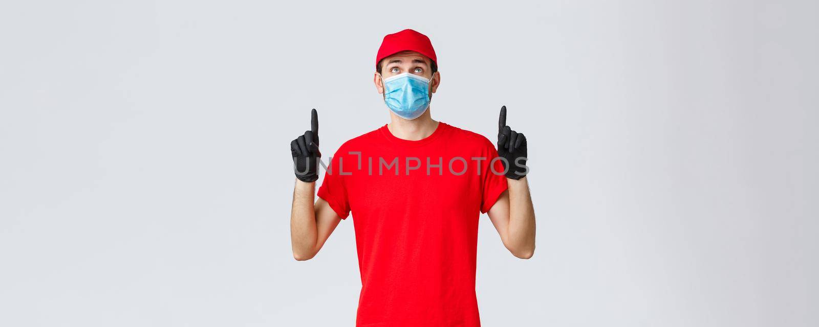 Covid-19, self-quarantine, online shopping and shipping concept. Curious delivery guy in red cap, t-shirt, medical mask and gloves, deliver self-quarantine orders to clients, looking pointing up.