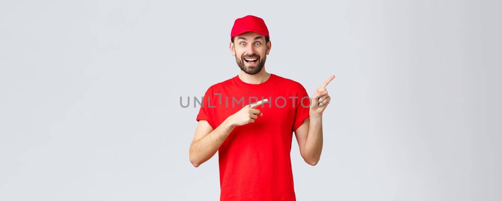 Online shopping, delivery during quarantine and takeaway concept. Enthusiastic smiling courier in red uniform cap and t-shirt, pointing fingers right, showing banner, recommend special offer by Benzoix