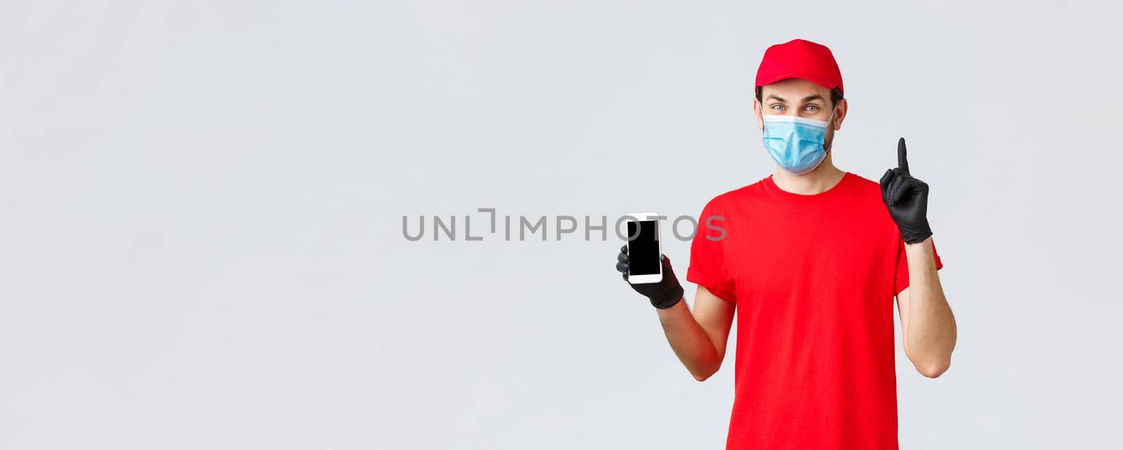 Contactless delivery, payment and online shopping during covid-19, self-quarantine. Friendly courier in red uniform, mask and gloves, pointing finger up, show smartphone display, order in internet by Benzoix