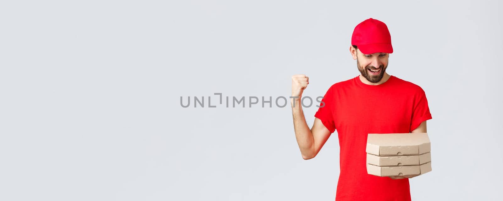 Food delivery, quarantine, stay home and order online concept. Happy, excited bearded courier in red t-shirt and cap celebrating, rejoicing, looking at pizza orders, grey background by Benzoix