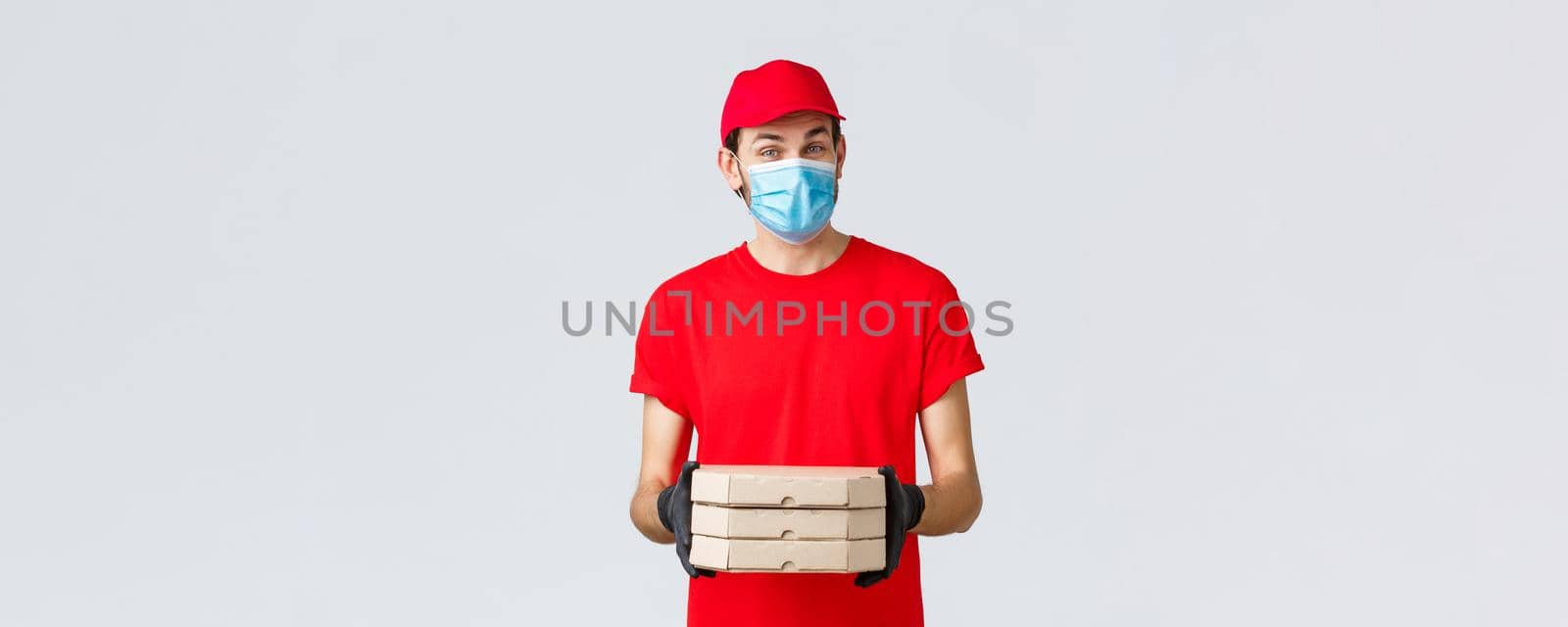 Food delivery, application, online grocery, contactless shopping and covid-19 concept. Pleasant courier in red uniform bring pizza in boxes, wear face mask and rubber gloves, give clint order by Benzoix