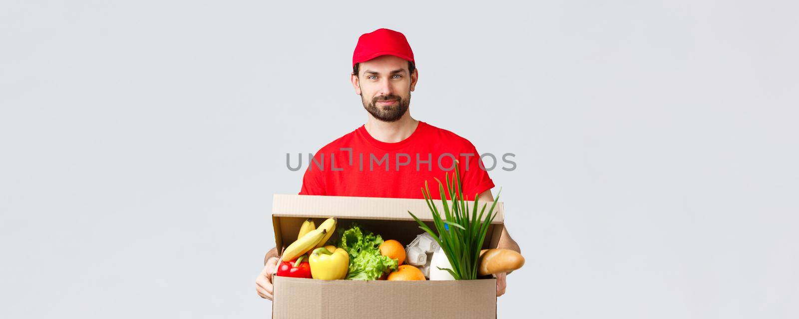 Groceries and packages delivery, covid-19, quarantine and shopping concept. Handsome courier in red uniform cap and t-shirt, bring grocery package, box delivery to client house, fast shipping by Benzoix