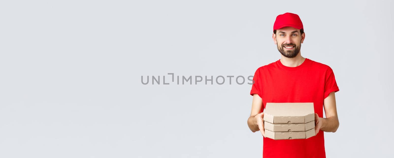 Food delivery, quarantine, stay home and order online concept. Smiling nice bearded courier in red uniform cap and t-shirt, handing clients boxes with pizza, deliver order, grey background.