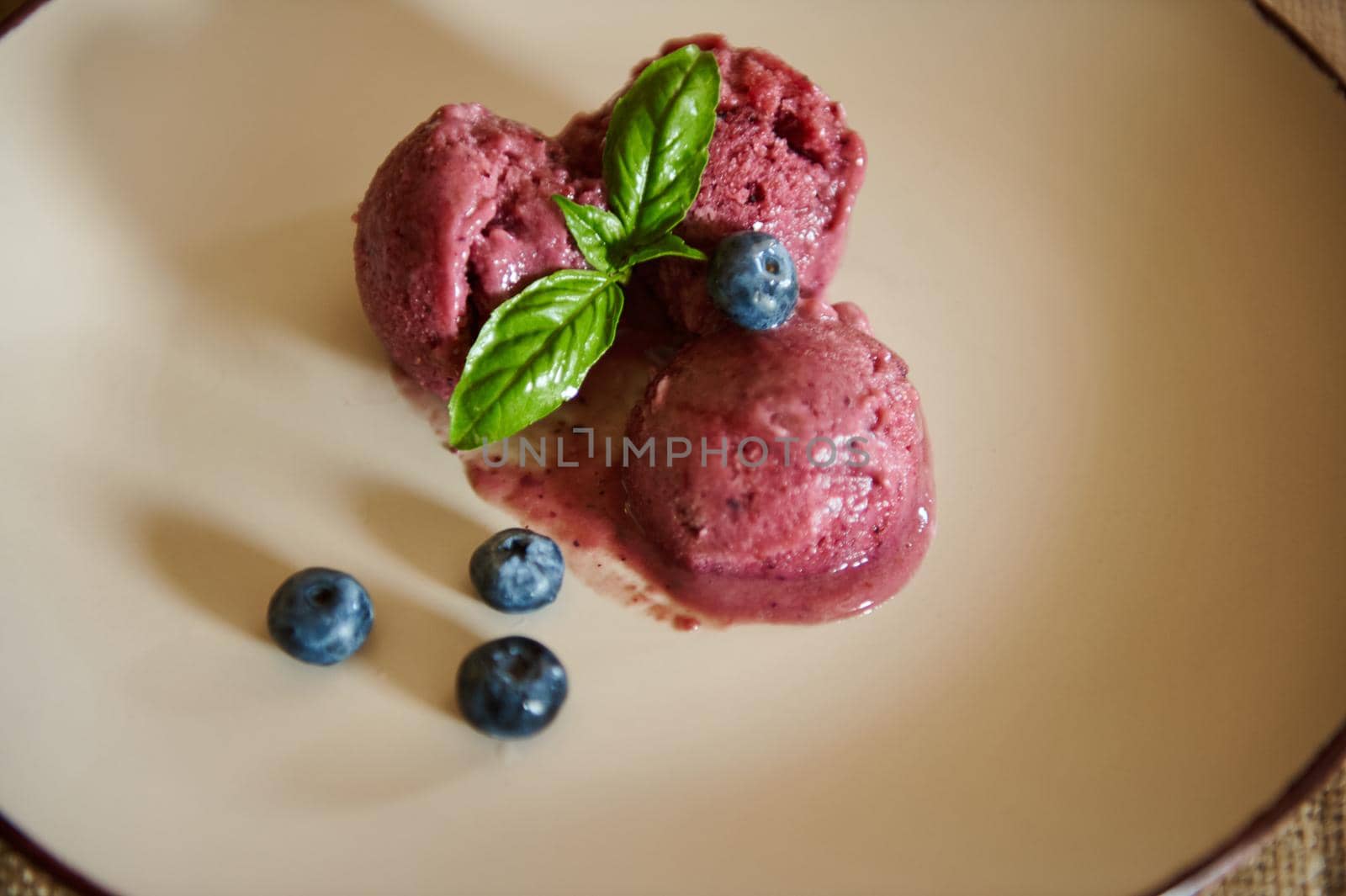 Still life. Close-up. Food consumerism. Three blueberry ice cream sorbet balls on a plate, decorated with blueberries and green leaves of a lemon basil and mint. Healthy frozen vegan sweet dessert