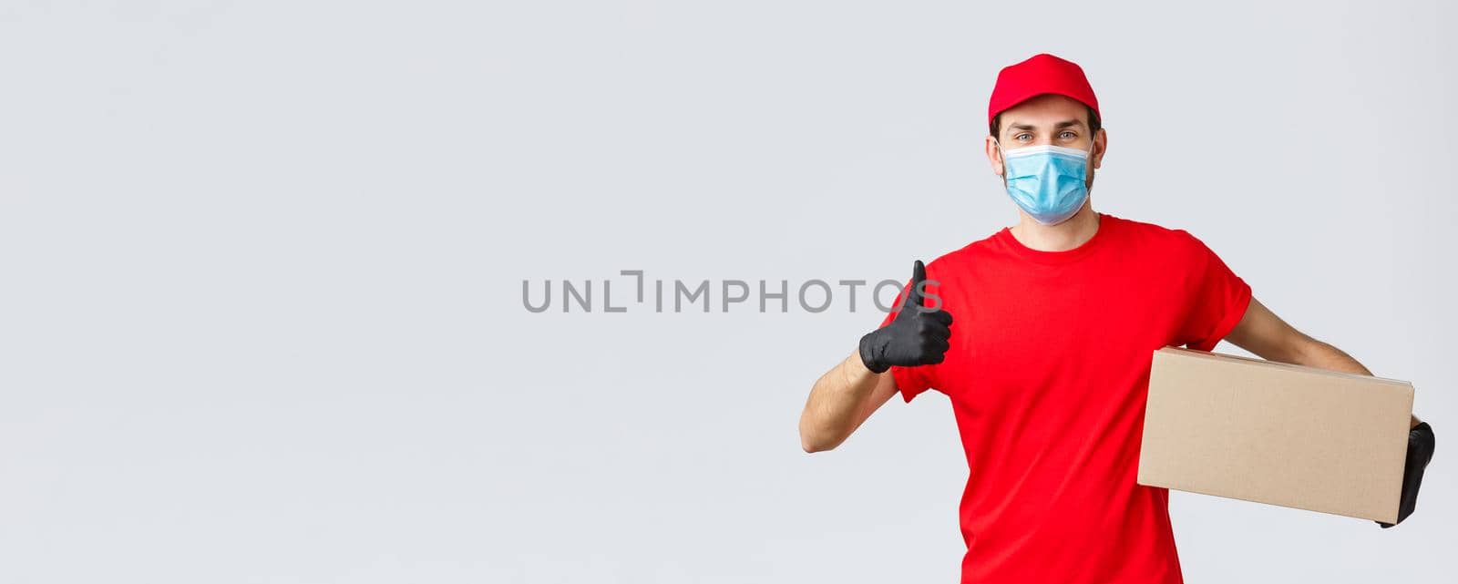 Packages and parcels delivery, covid-19 quarantine delivery, transfer orders. Cheerful courier in red uniform, gloves and face mask, thumb-up, recommend contactless deliver, holding box with order.