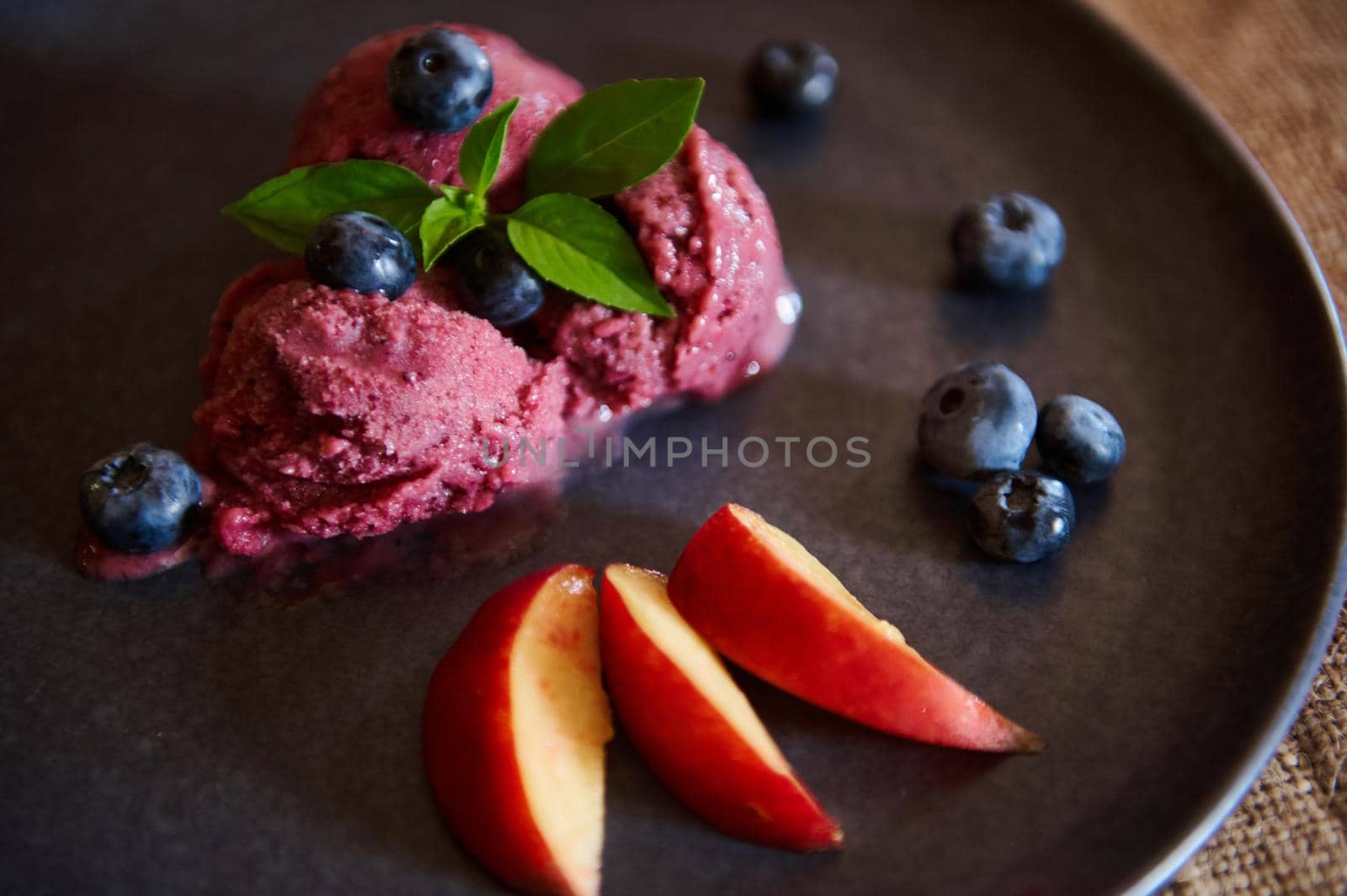 Still life of a healthy, sweet, raw vegan dessert. Homemade blueberry sorbet, ice cream with slices of ripe juicy peach and lemon basil leaves on a navy color ceramic plate. Food and drink consumerism