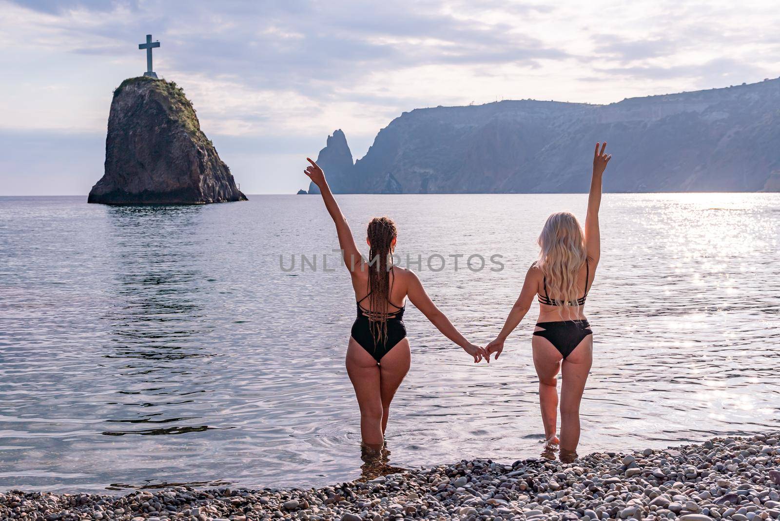 Two beautiful young female friends in black bikinis opened their arms in delight in front of the blue sea and sky.
