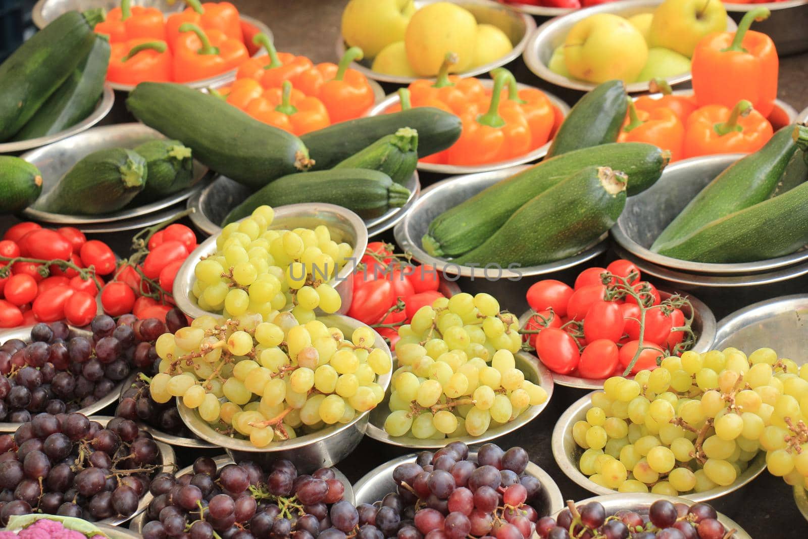 Fresh fruits and vegetables on a market stall: grapes, tomatoes, courgette and peppers displayed in small metal bowls