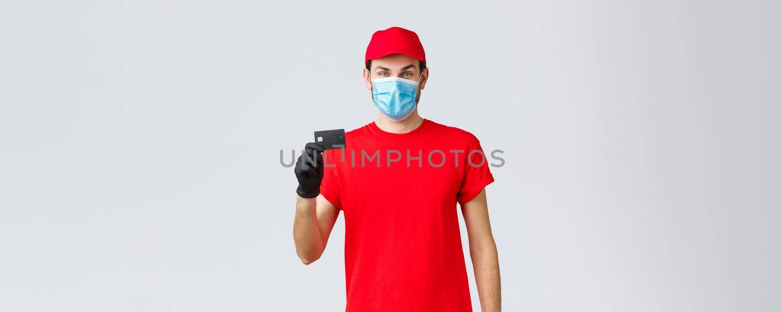 Contactless delivery, payment and online shopping during covid-19, self-quarantine. Young courier in red uniform cap, face mask and gloves, showing credit card, easy paying and ordering by Benzoix