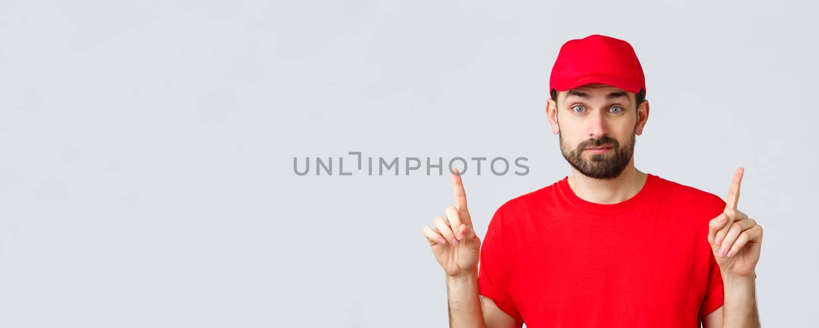 Online shopping, delivery during quarantine and takeaway concept. Indecisive and unsure courier in red uniform cap, t-shirt, smirk and pointing fingers up uncertain, dont know, cant decide by Benzoix