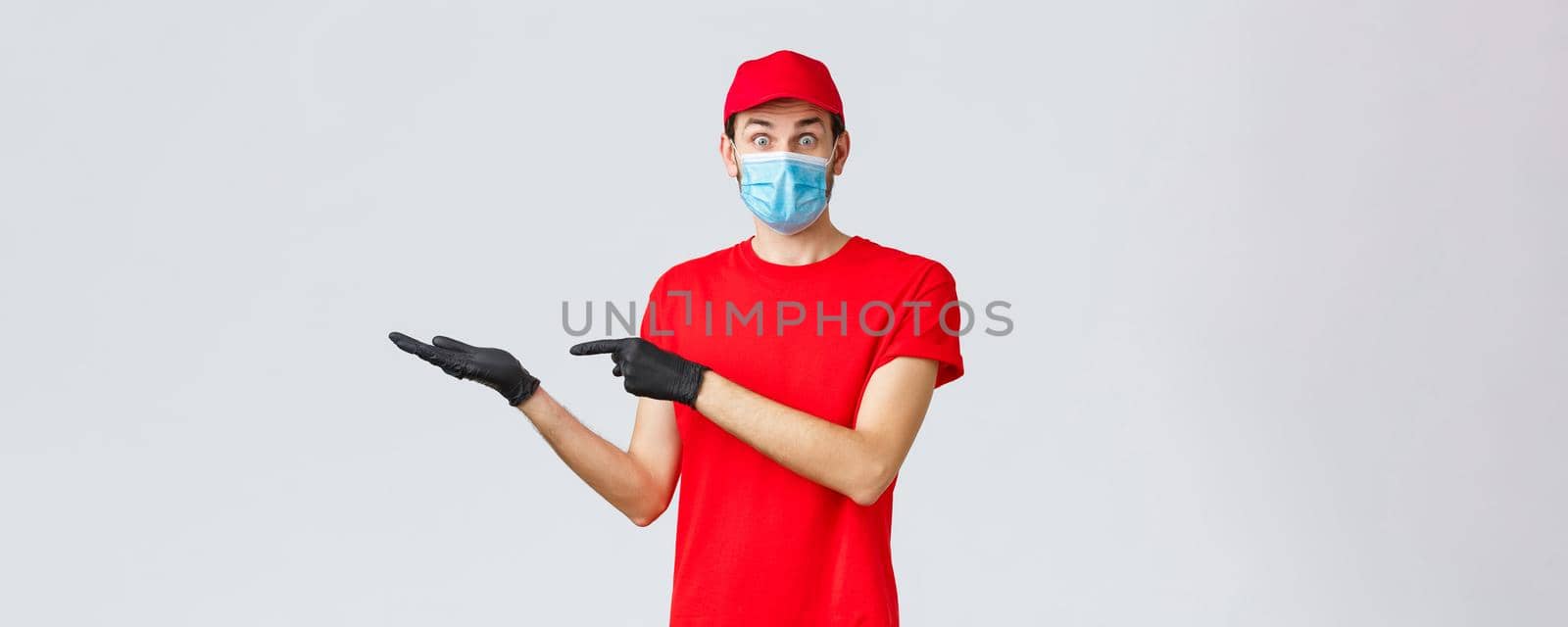 Groceries and packages delivery, covid-19, quarantine and shopping concept. Surprised and excited courier in red uniform, face mask and gloves introduce good new bonus offer, promo, showing banner by Benzoix