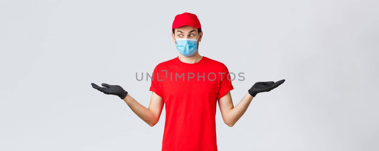 Groceries and packages delivery, covid-19, quarantine and shopping concept. Surprised courier in red cap and t-shirt uniform, wear protective face mask, gloves, hold two items in hands sideways.