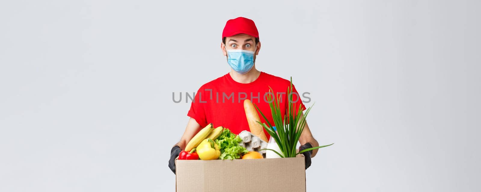 Groceries and packages delivery, covid-19, quarantine and shopping concept. Serious courier in red uniform, gloves and protective face mask, deliver package box to client house during coronavirus.