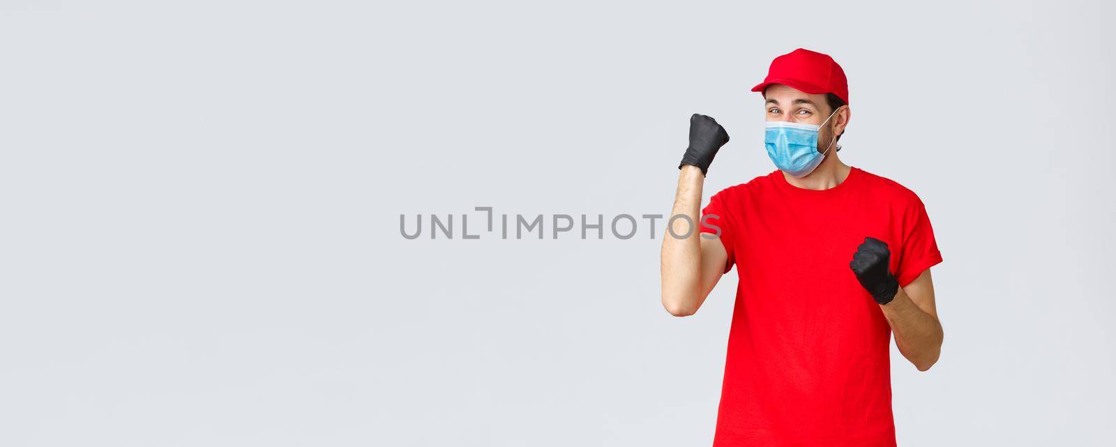Covid-19, self-quarantine, online shopping and shipping concept. Cheerful delivery man in red uniform, gloves and face mask, do champion dance, celebrating success or achievement, courier fist pump by Benzoix