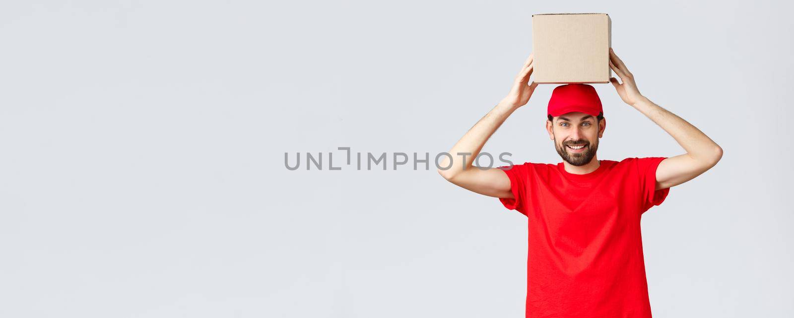 Order delivery, online shopping and package shipping concept. Funny and cute bearded courier in red uniform cap and t-shirt, holding box on head. Employee with package smiling.