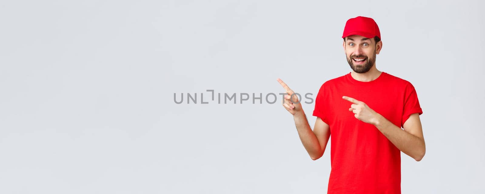 Online shopping, delivery during quarantine and takeaway concept. Amused and excited smiling courier inviting client take look, employee in red uniform cap and t-shirt pointing fingers left.