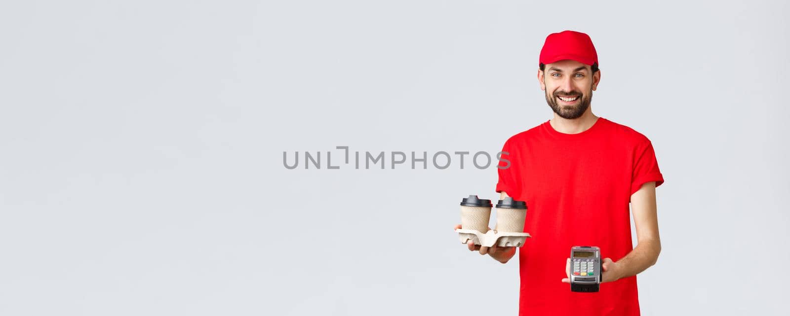 Food delivery, quarantine, stay home and order online concept. Friendly bearded courier in red uniform handing coffee delivery and POS terminal for client to pay contactless, grey background.