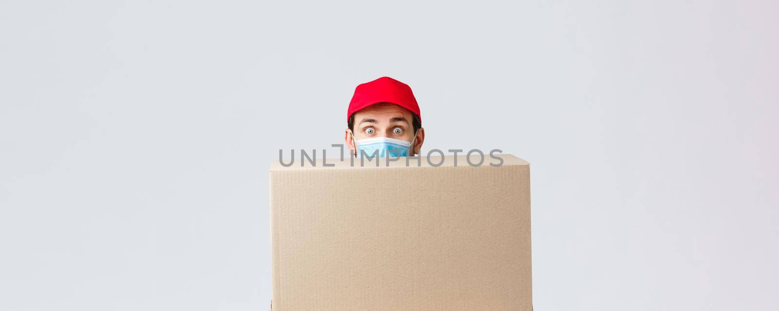 Packages and parcels delivery, covid-19 quarantine and transfer orders. Clumsy cute courier first day at job, hiding behind large box, wear face mask and red uniform cap, grey background by Benzoix