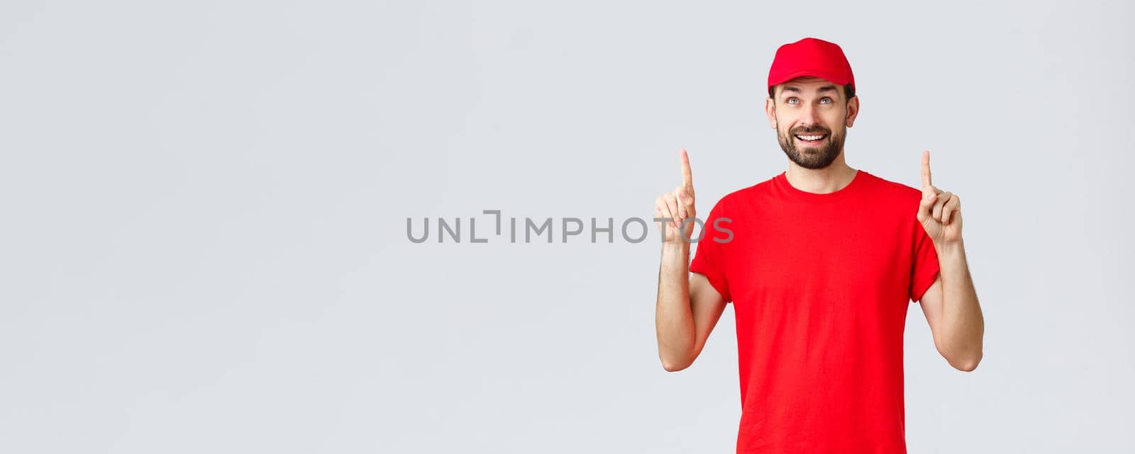 Online shopping, delivery during quarantine and takeaway concept. Smiling cheerful courier in red t-shirt, cap pointing fingers up. Employee look intrigued at banner with special discount by Benzoix