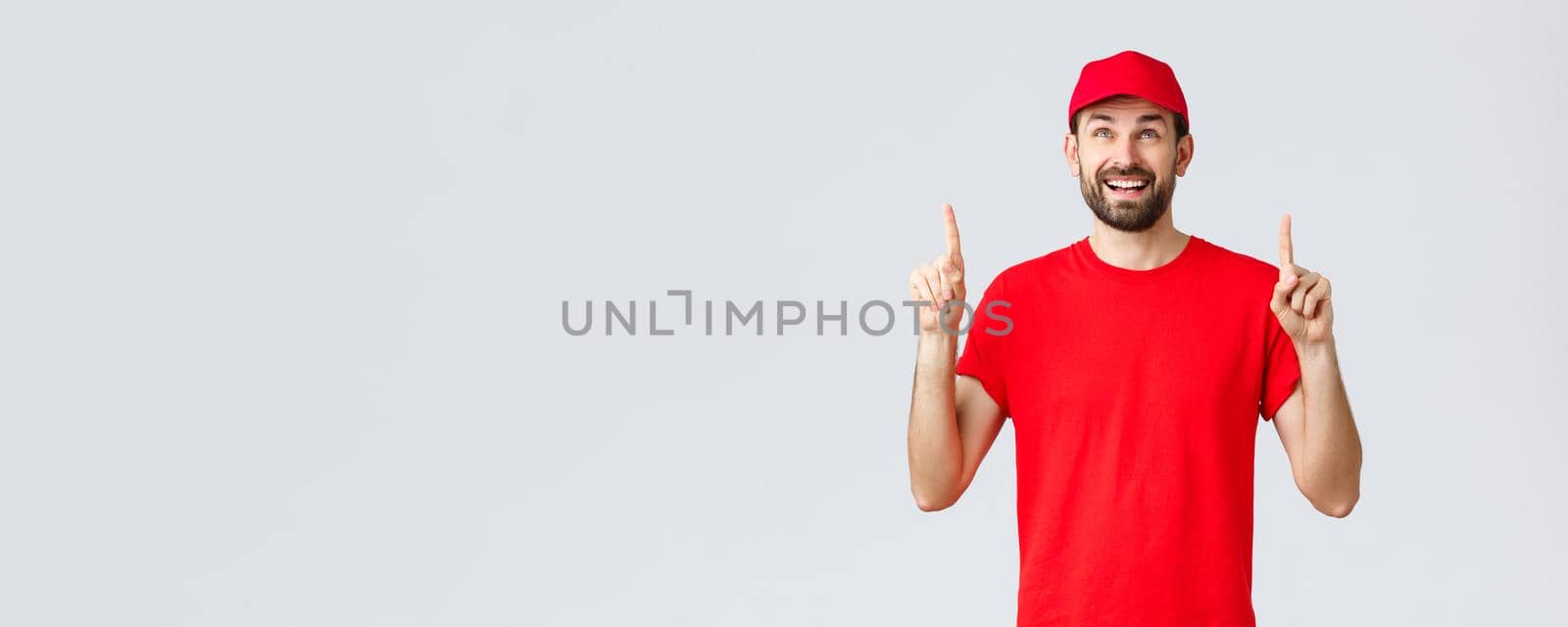 Online shopping, delivery during quarantine and takeaway concept. Cheerful excited courier in red uniform cap and t-shirt, smiling amazed and pointing fingers up, reading banner or sign.