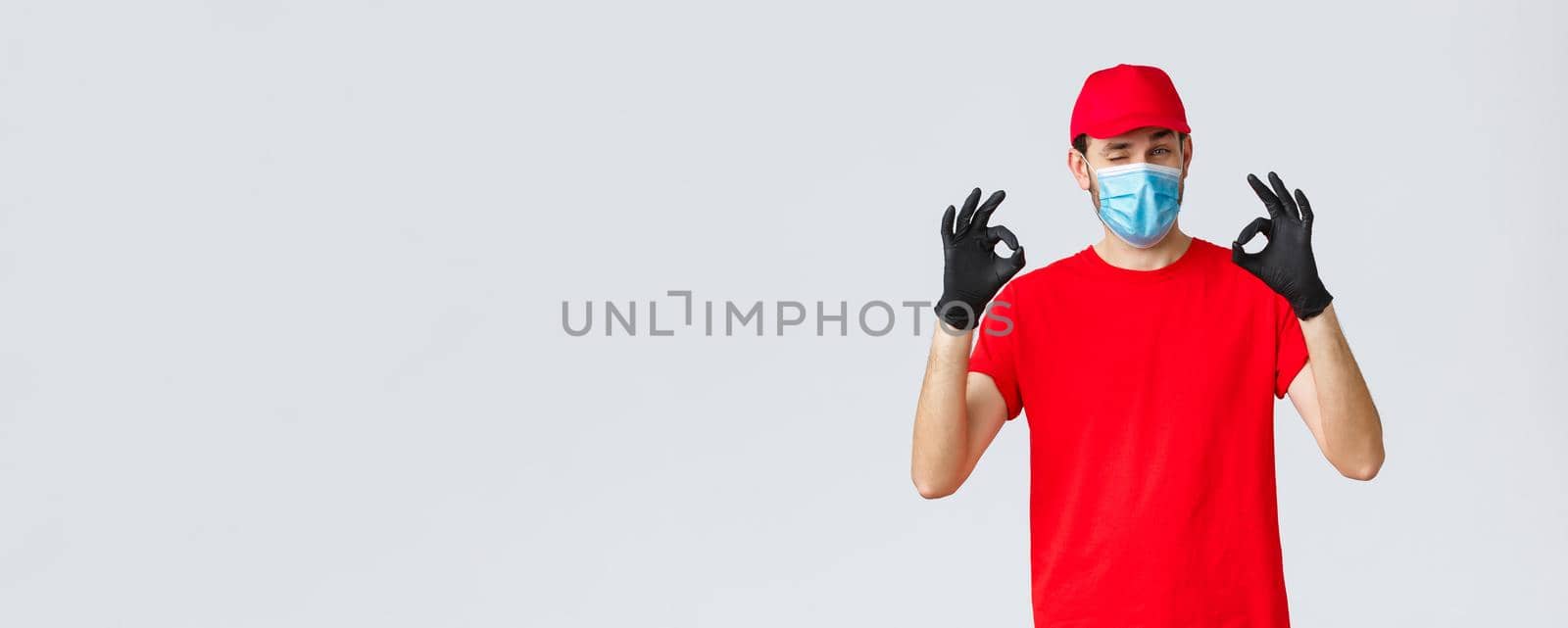 Covid-19, self-quarantine, online shopping and shipping concept. Handsome delivery guy in red cap, t-shirt, wear protective face mask and gloves from coronavirus, show okay, agree and good gesture.