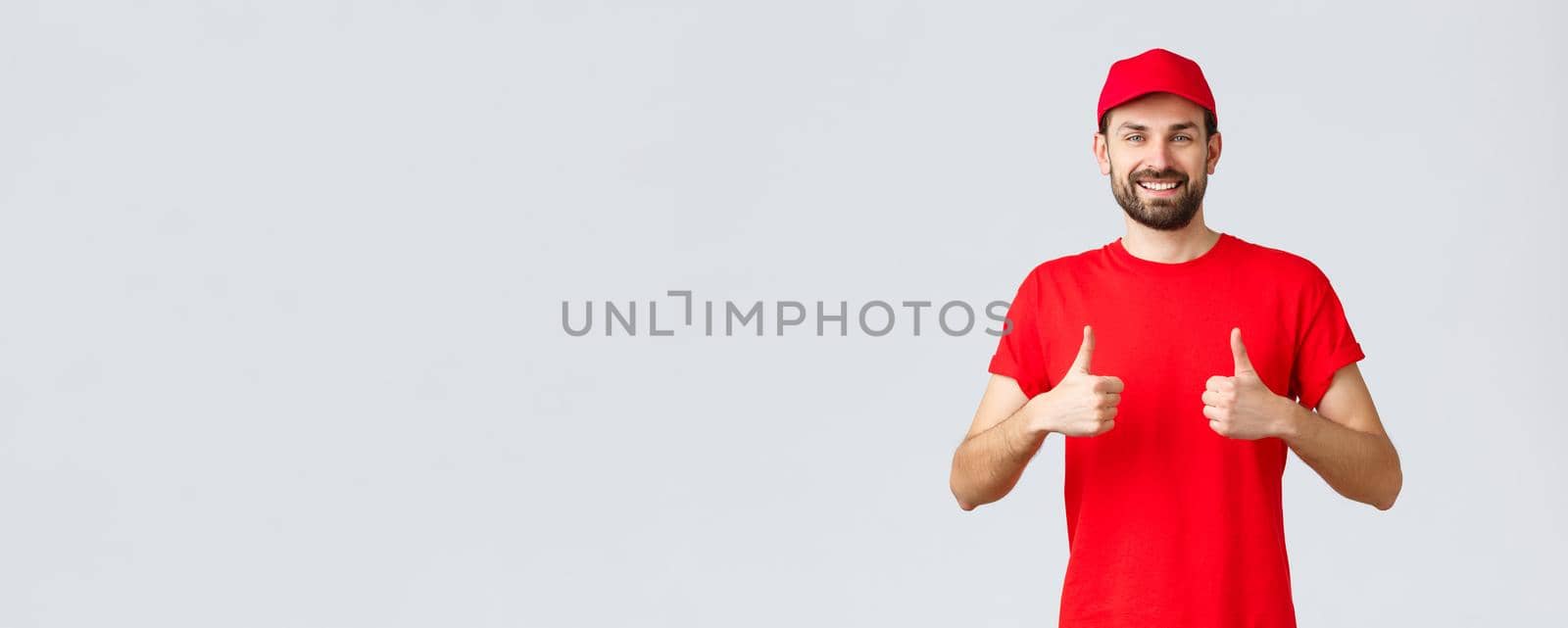Online shopping, delivery during quarantine and takeaway concept. Cheerful courier in red uniform cap and t-shirt, recommends make orders, thumbs-up in approval, grey background.
