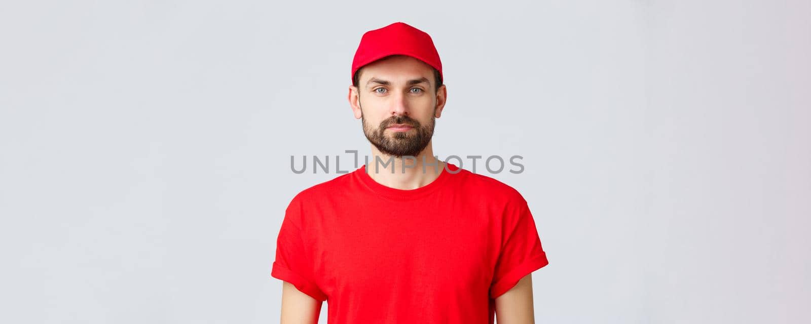 Online shopping, delivery during quarantine and takeaway concept. Young handsome bearded courier in red service uniform with cap and t-shirt, employee working transfering orders.