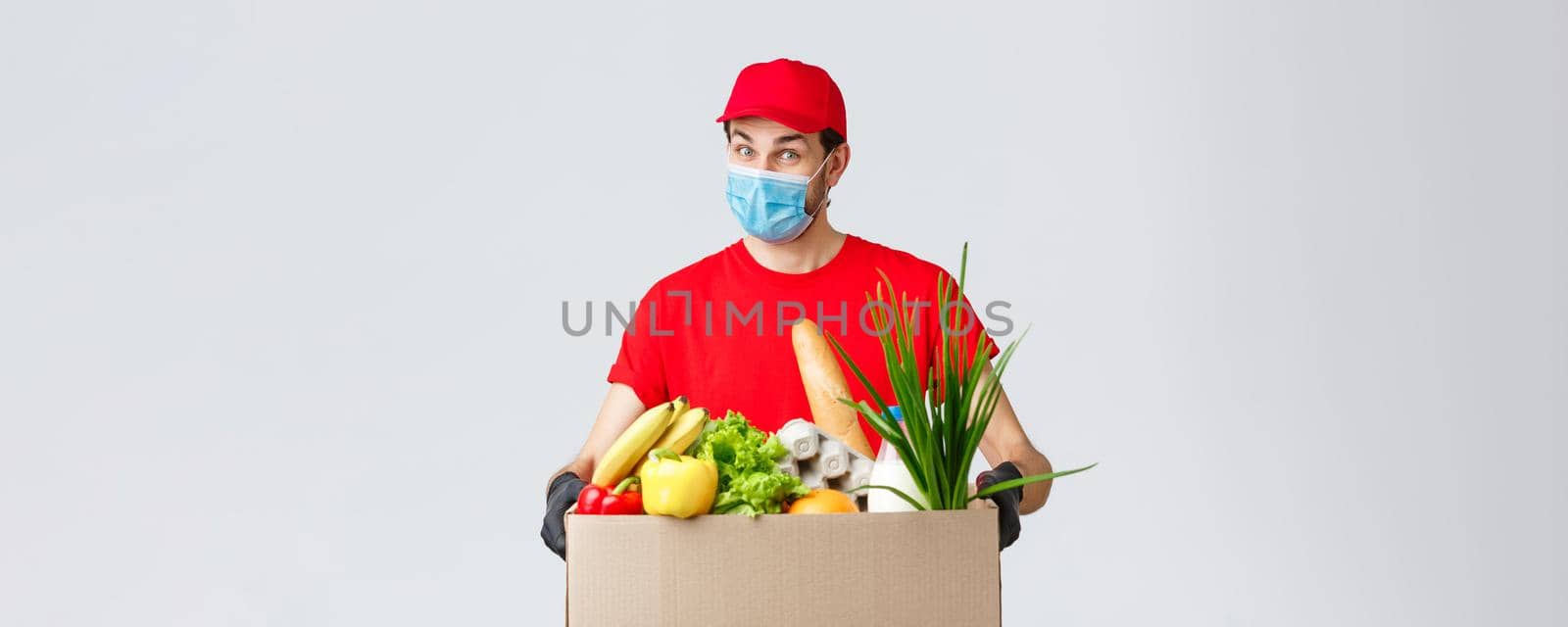 Groceries and packages delivery, covid-19, quarantine and shopping concept. Friendly courier in face mask and gloves, red uniform bring food box to customer ordered online, contactless deliver.