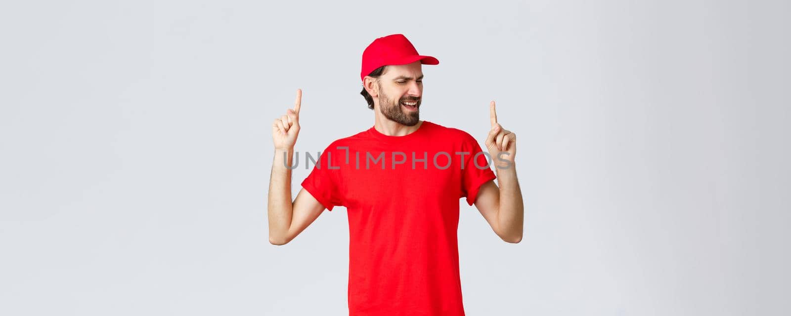 Upbeat handsome bearded delivery guy spending break relaxing, employee in red uniform cap and t-shirt singing song, pointing fingers up carefree, close eyes, standing grey background by Benzoix