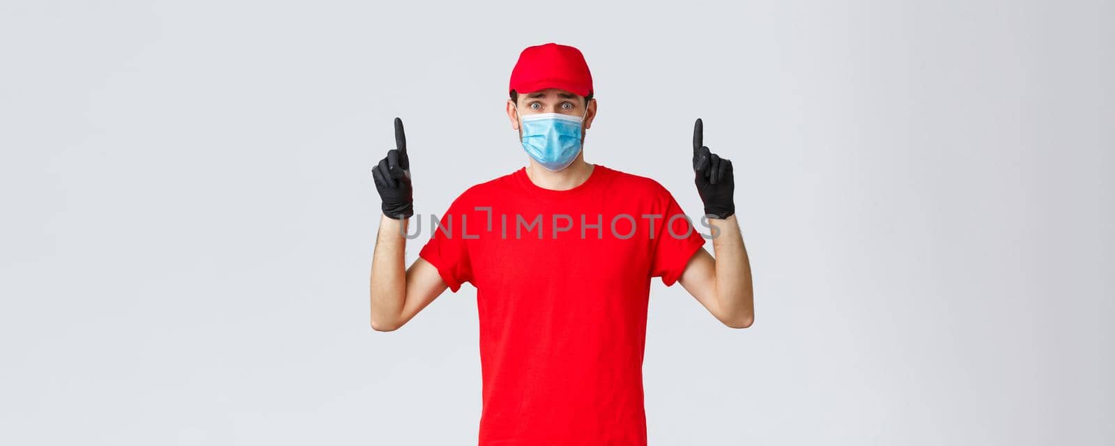Covid-19, self-quarantine, online shopping and shipping concept. Nervous or scared, worried delivery man in red uniform cap, t-shirt, frowning pointing fingers up at bad news, wear mask and gloves by Benzoix