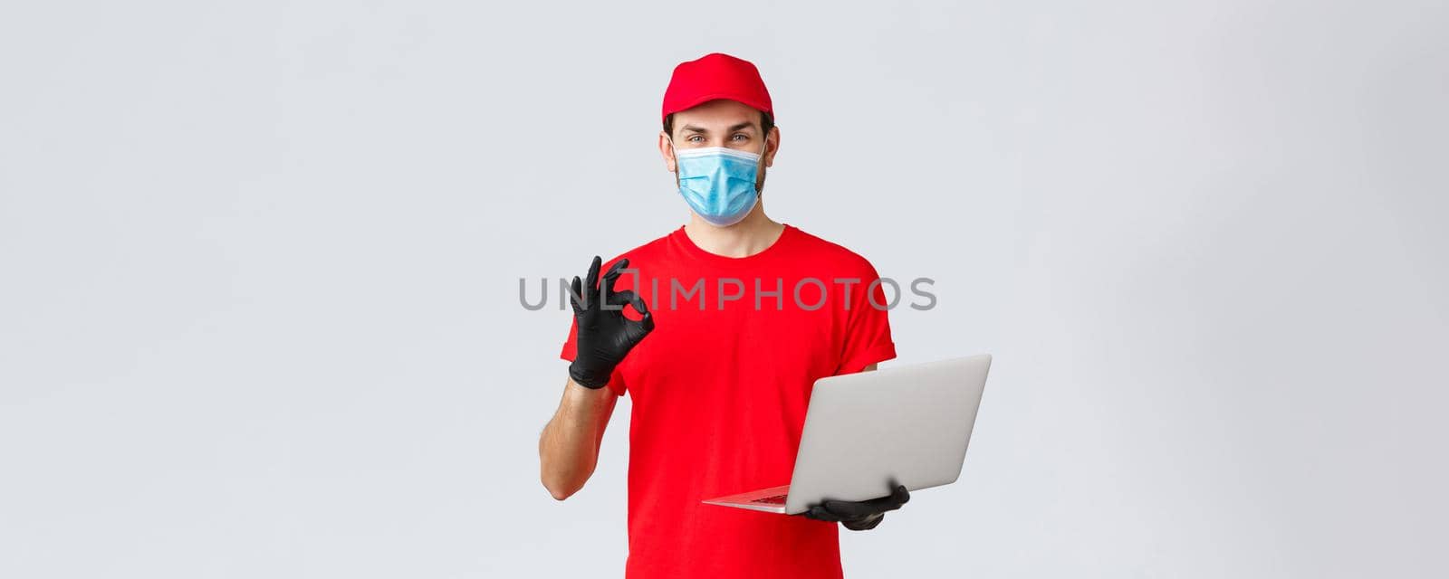 Customer support, covid-19 delivery packages, online orders processing concept. Smiling courier in face mask and gloves guarantee safety of parcel, processing order, show okay sign, hold laptop.