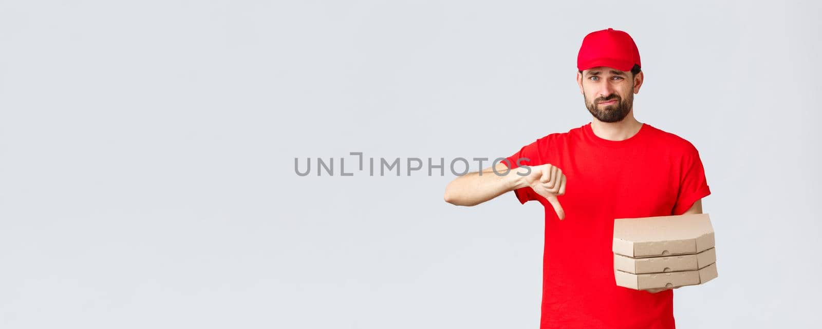 Food delivery, quarantine, stay home and order online concept. Disappointed and reluctant courier in red uniform cap and t-shirt, dislike pooq quality fast-food, thumb-down, holding pizza by Benzoix