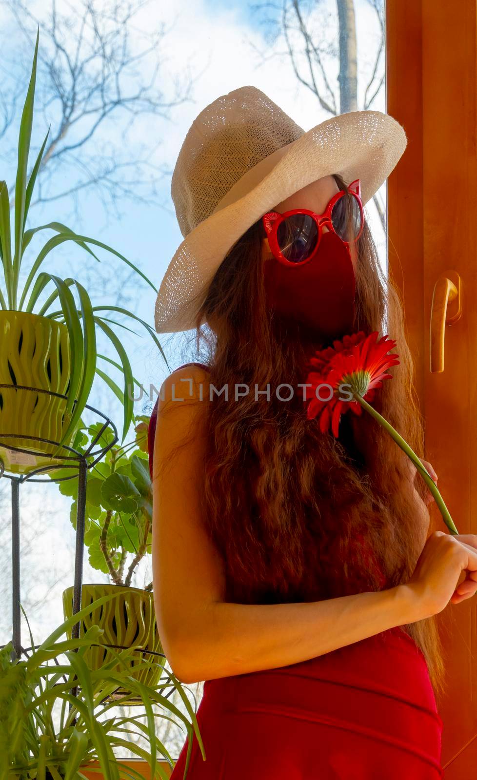 A girl in a red dress, a white hat, funny sunglasses and a red medical mask is hold red flower with plants against the sky in early sprin