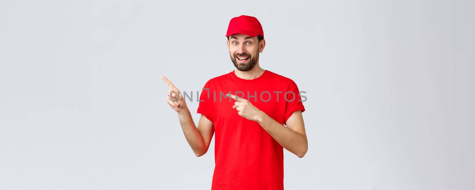 Online shopping, delivery during quarantine and takeaway concept. Amused and excited smiling courier inviting client take look, employee in red uniform cap and t-shirt pointing fingers left.