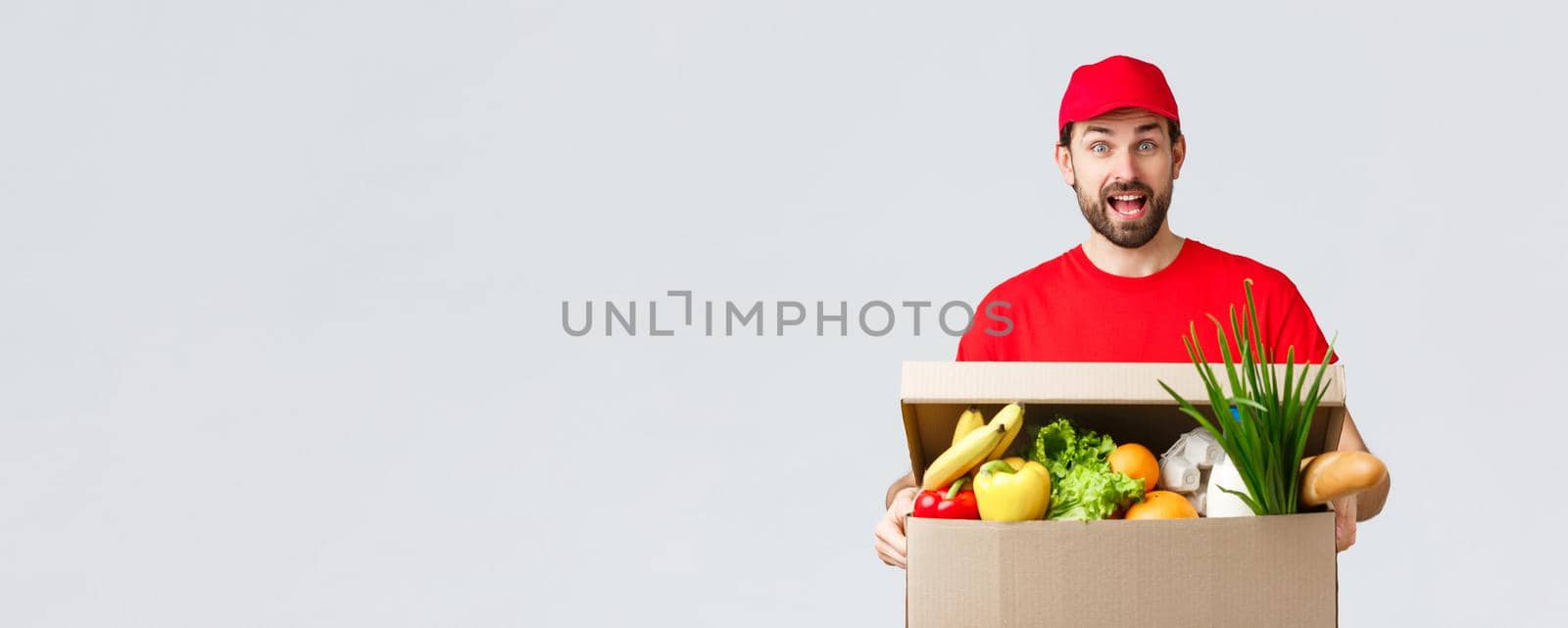 Groceries and packages delivery, covid-19, quarantine and shopping concept. Smiling handsome bearded courier in red uniform, bring food package, grocery order to client in box, look amused.