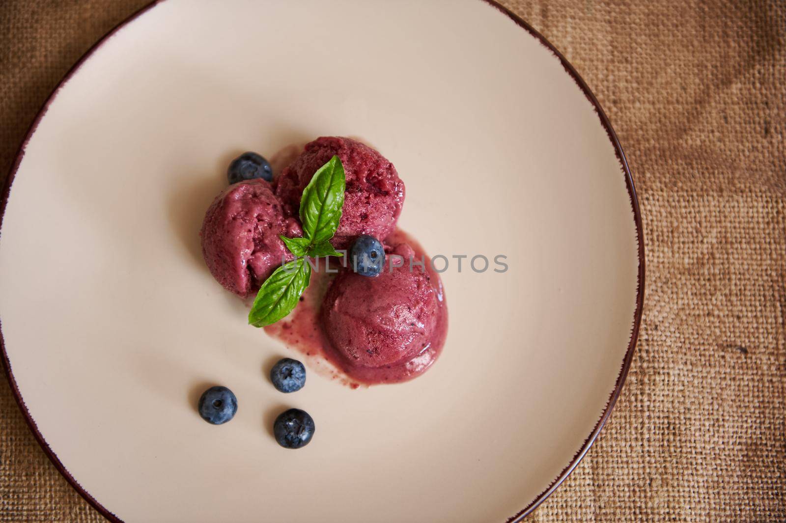 Flat lay. Tasty, vegan sorbet. Homemade huckleberry ice cream, topped with organic blueberries and green leaf of lemon basil. Tree frozen balls of banana and berry on a beige plate. Healthy eating