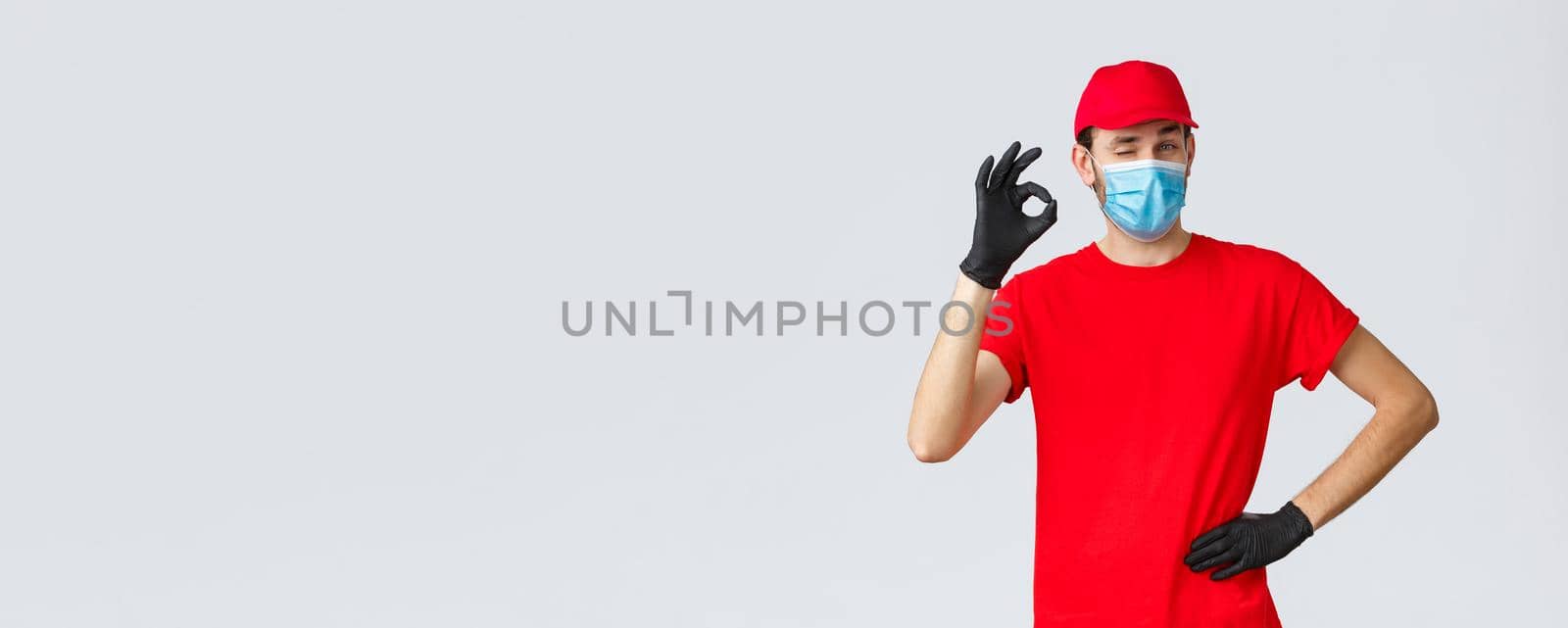 Covid-19, self-quarantine, online shopping and shipping concept. Delivery guy in red t-shirt, cap with face mask and gloves, show okay sign, wink, say no problem. Courier deal with any transfer by Benzoix