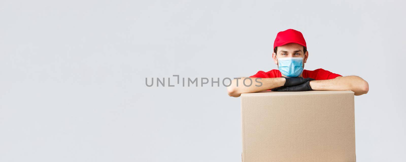 Packages and parcels delivery, covid-19 quarantine and transfer orders. Young courier in red uniform cap, face mask and gloves, leaning on box to deliver, shipping your orders, grey background by Benzoix