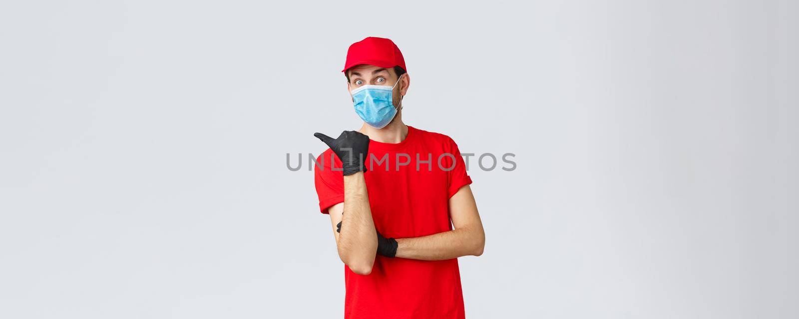 Covid-19, self-quarantine, online shopping and shipping concept. Excited surprised delivery man in red uniform, face mask and gloves, pointing left at promo, showing clients bonuses or application by Benzoix