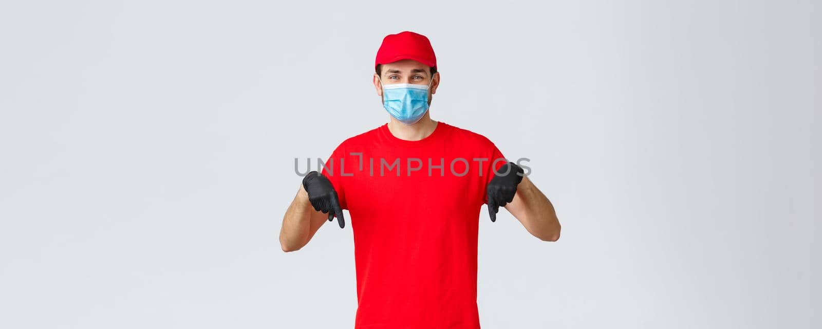 Covid-19, self-quarantine, online shopping and shipping concept. Smiling friendly delivery guy red uniform, cap and t-shirt, wear medical mask with gloves, pointing down, showing promo or client info.