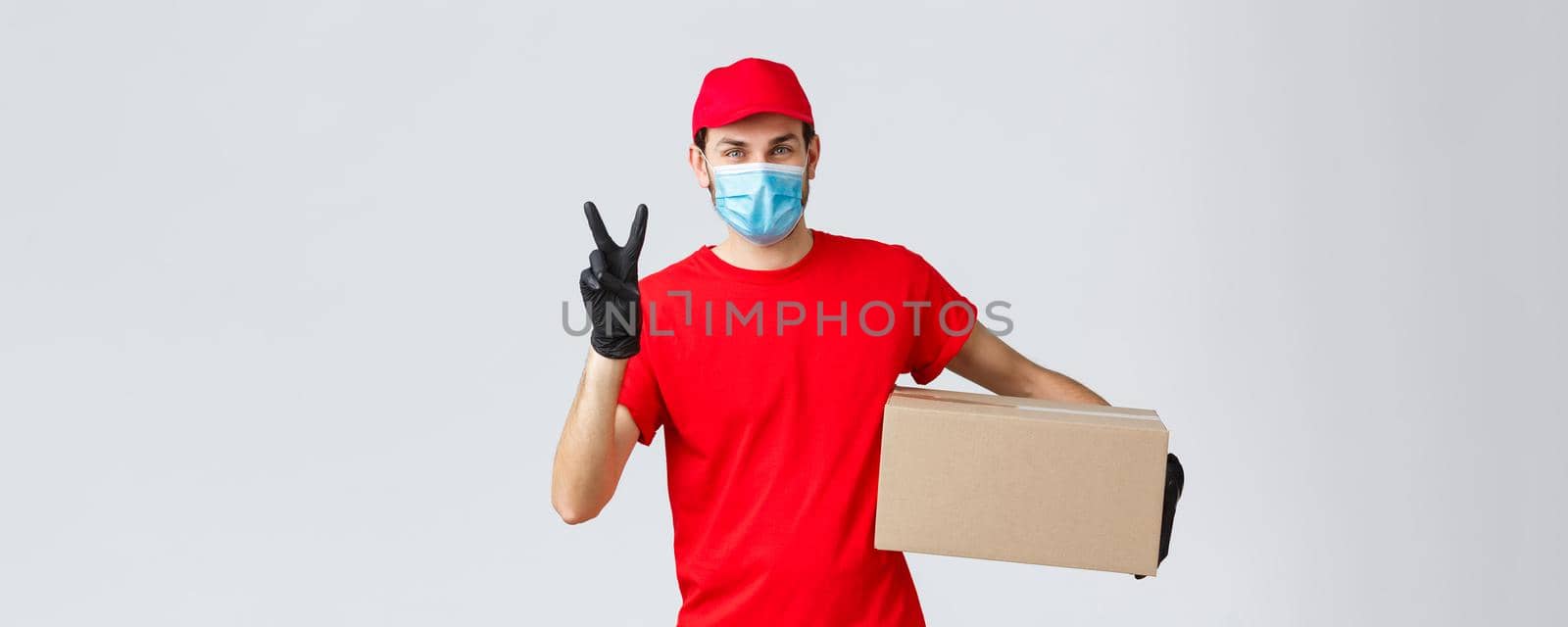 Packages and parcels delivery, covid-19 quarantine delivery, transfer orders. Friendly courier in red uniform, face mask and gloves, deliver order to client, holding box, show peace sign.