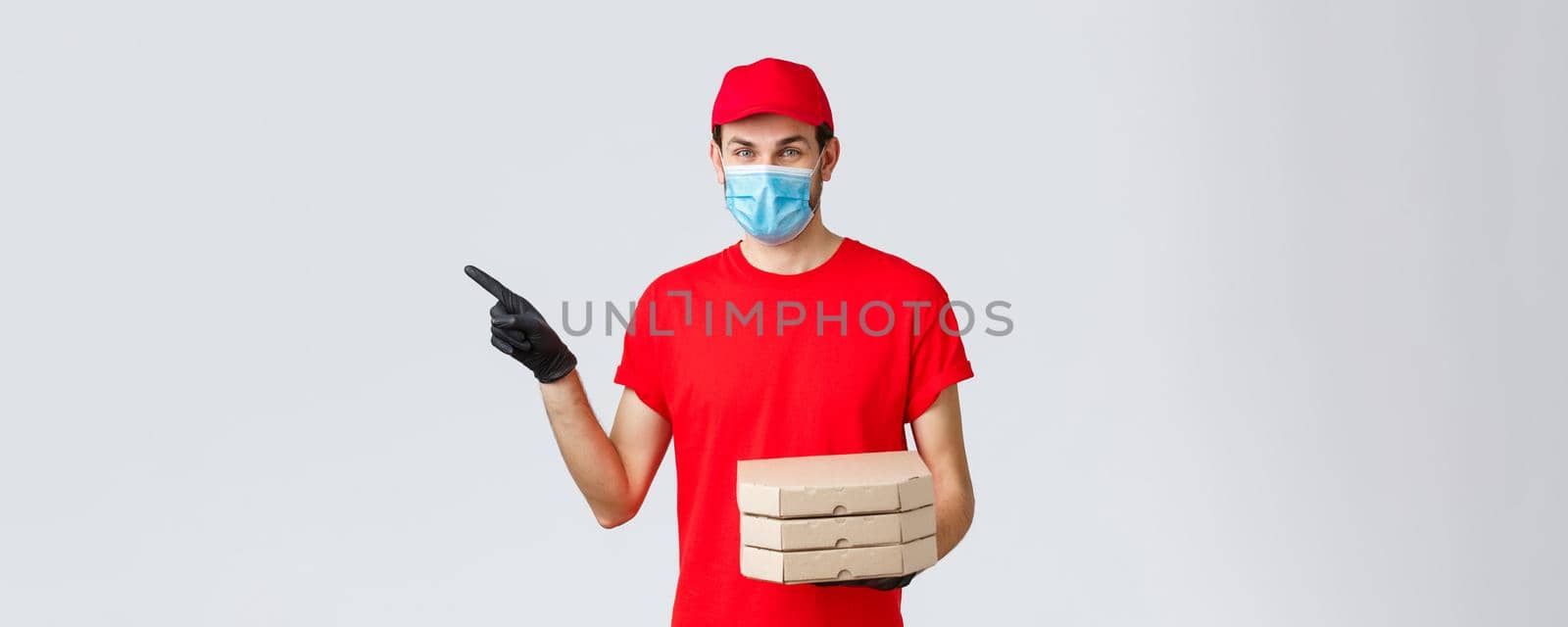 Food delivery, application, online contactless shopping and covid-19 concept. Charismatic courier in red uniform, face mask and gloves, pointing finger left, holding pizza, bring order client.
