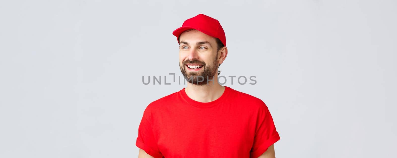 Online shopping, delivery during quarantine and takeaway concept. Cheerful bearded guy in red uniform cap and t-shirt, looking away with pleased smile, reading banner sign, grey background.
