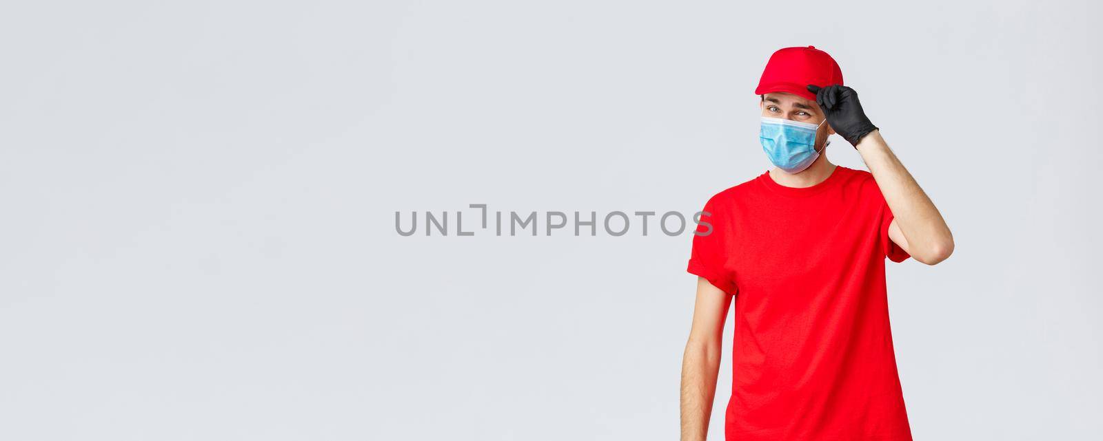 Covid-19, self-quarantine, online shopping and shipping concept. Sassy delivery man in red uniform and mask with gloves, saluting client, deliver packages and orders during coronavirus, food orders by Benzoix