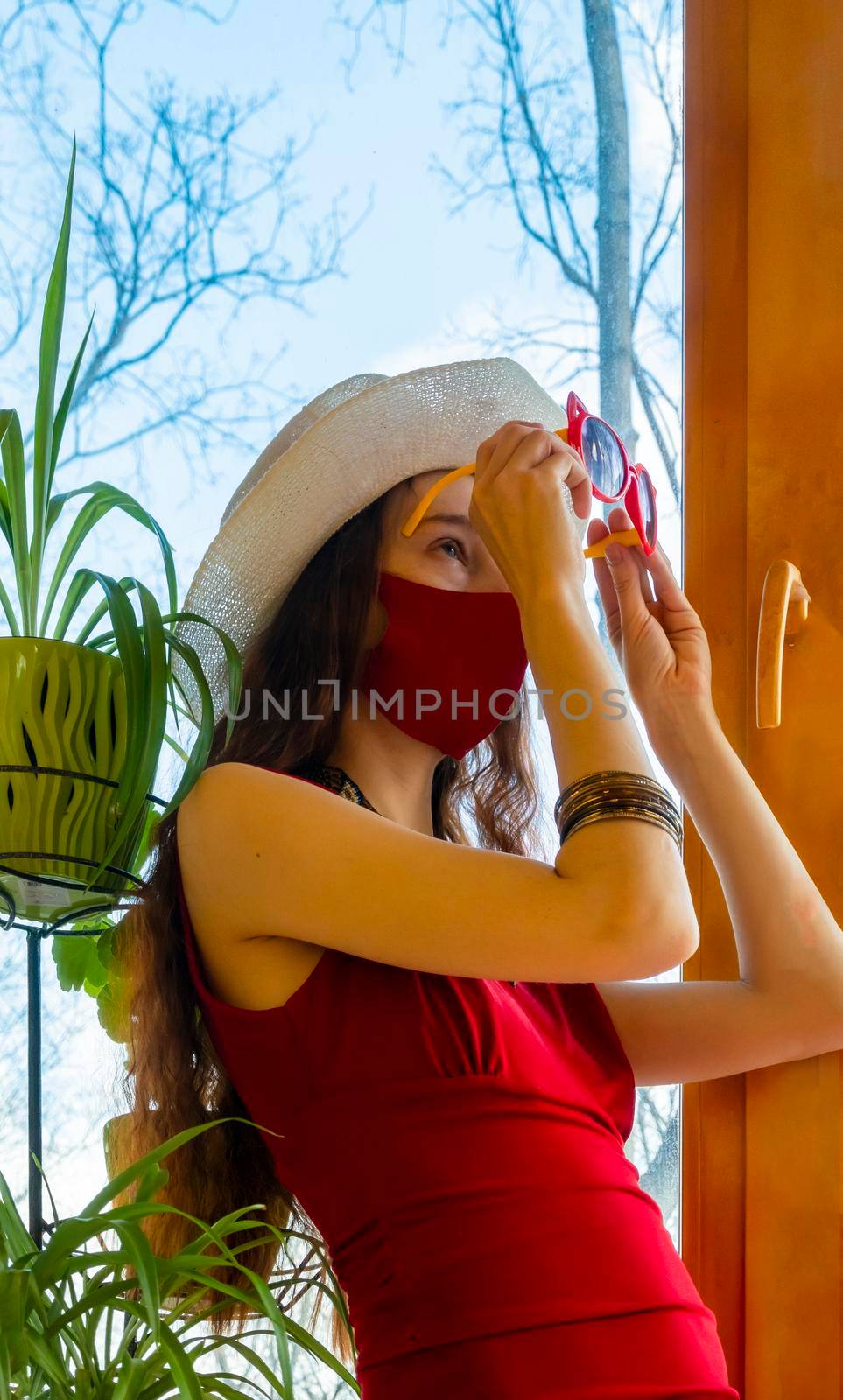 A girl in a red dress, a white hat and a red medical mask is wearing funny sunglasses with plants against the sky in early spring