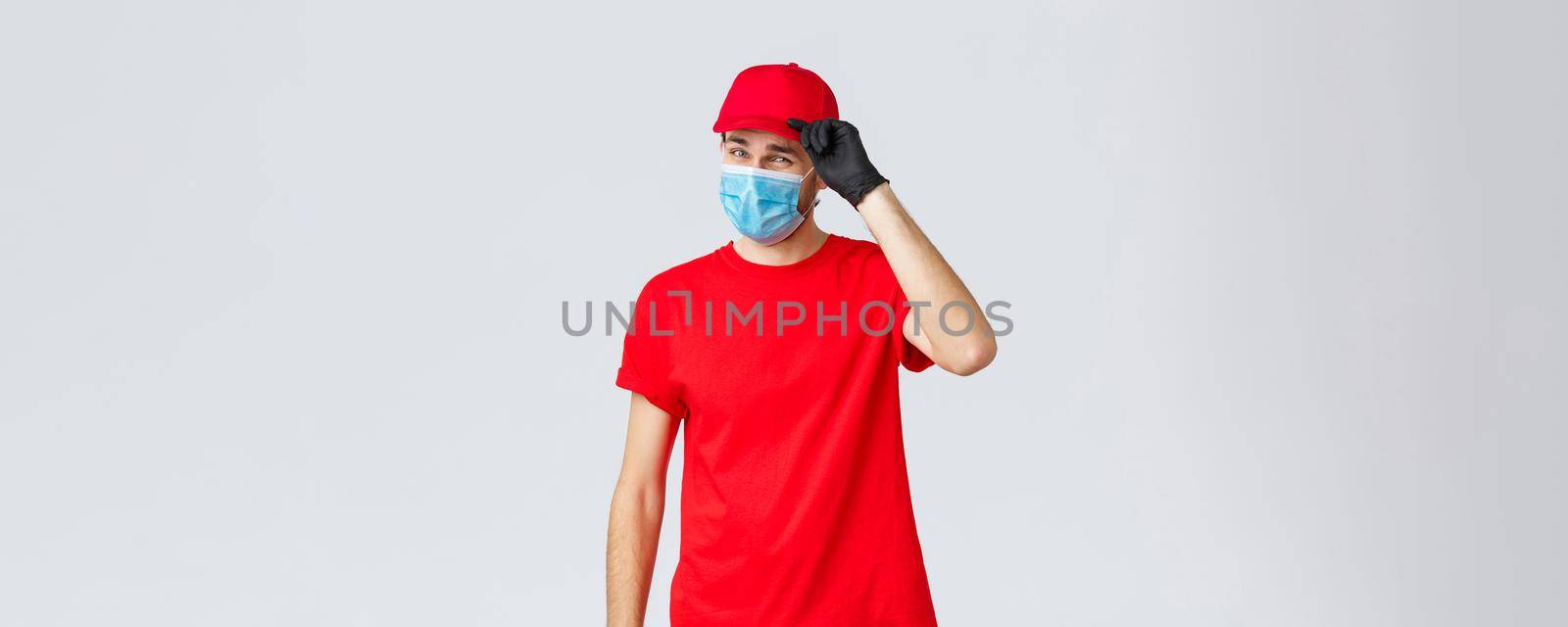 Covid-19, self-quarantine, online shopping and shipping concept. Sassy delivery man in red uniform and mask with gloves, saluting client, deliver packages and orders during coronavirus, food orders.