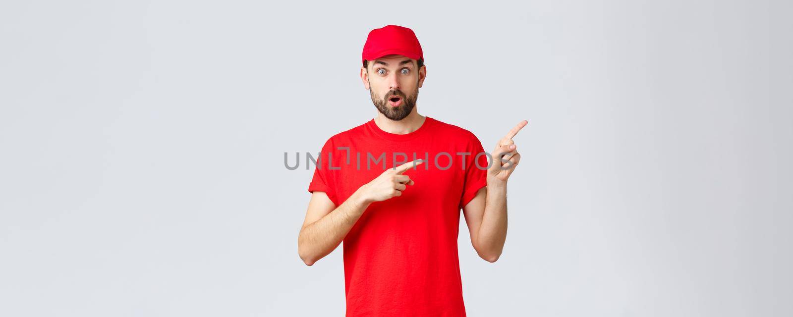 Online shopping, delivery during quarantine and takeaway concept. Take quick look here. Impressed courier showing clients special promo. Employee in red uniform pointing fingers right.