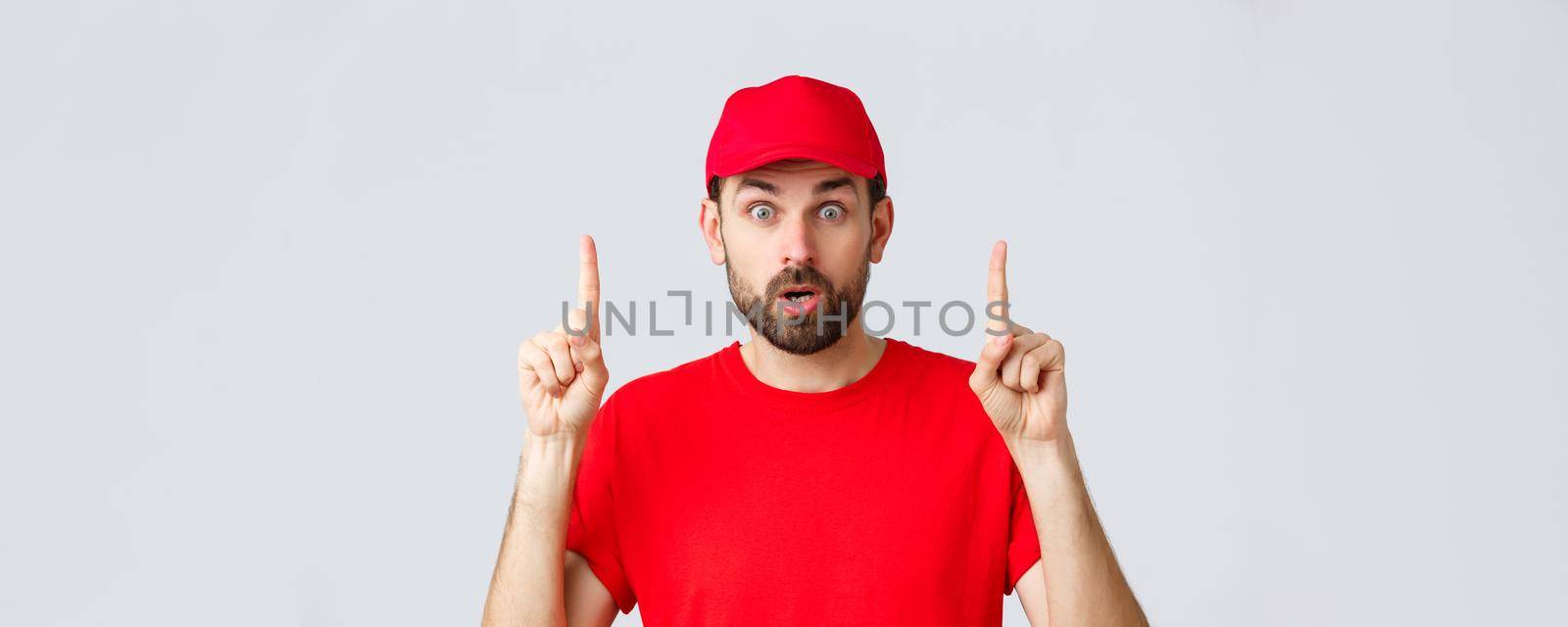 Online shopping, delivery during quarantine and takeaway concept. Surprised and impressed delivery guy, courier in red t-shirt and cap, pointing fingers up, gasping amazed, read interesting info by Benzoix