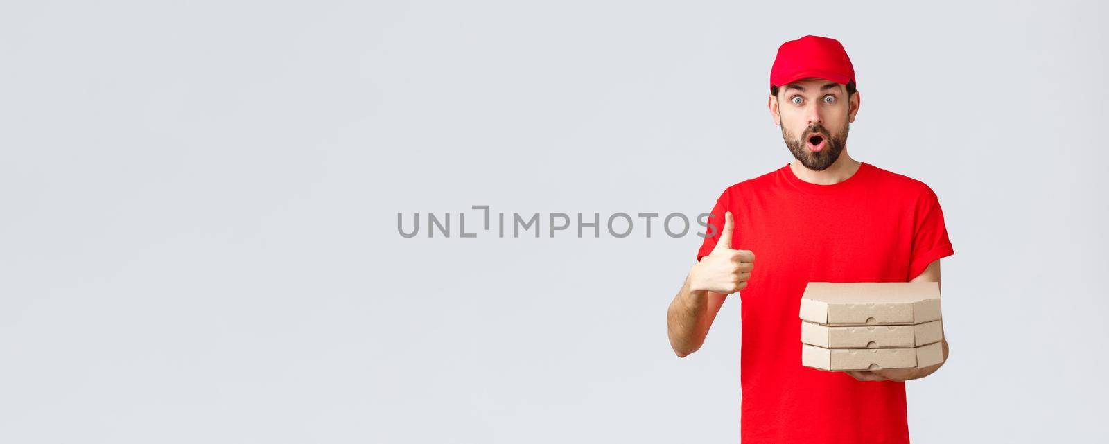 Food delivery, quarantine, stay home and order online concept. Surprised and amazed bearded courier in red t-shirt, cap gasping impressed, show thumb-up, bring order pizza, grey background.